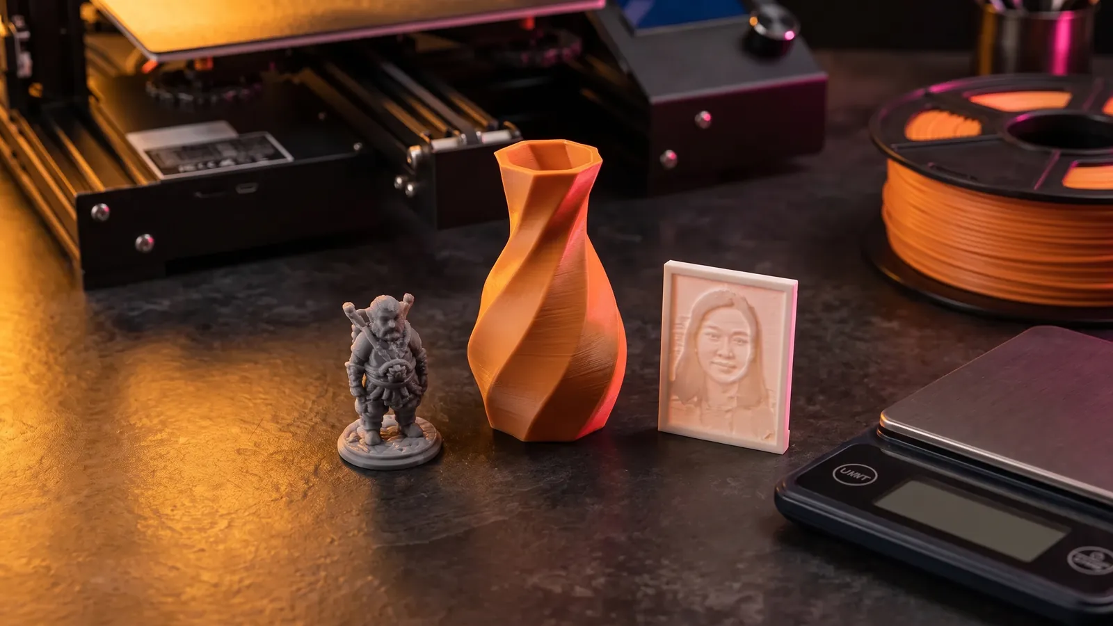 A grey printed figurine, an orange twisted vase and a small lithophane next to a filament spool and a kitchen scale on a dark workbench